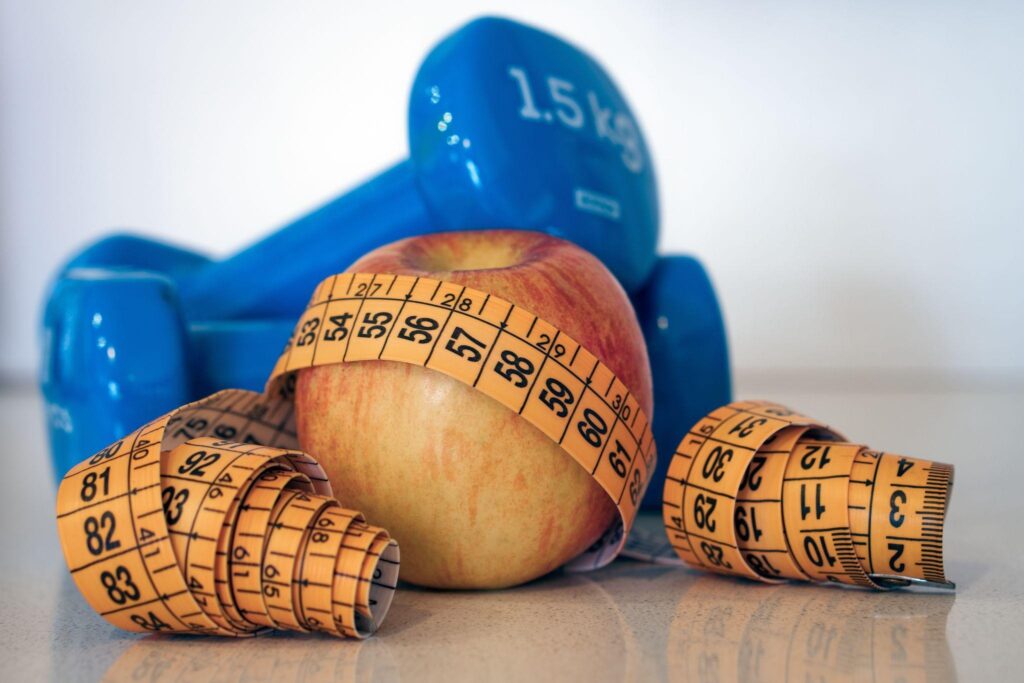 Apple wrapped in a measuring tape in front of blue 1.5 kg dumbbells, symbolizing healthy weight loss and fitness goals. 