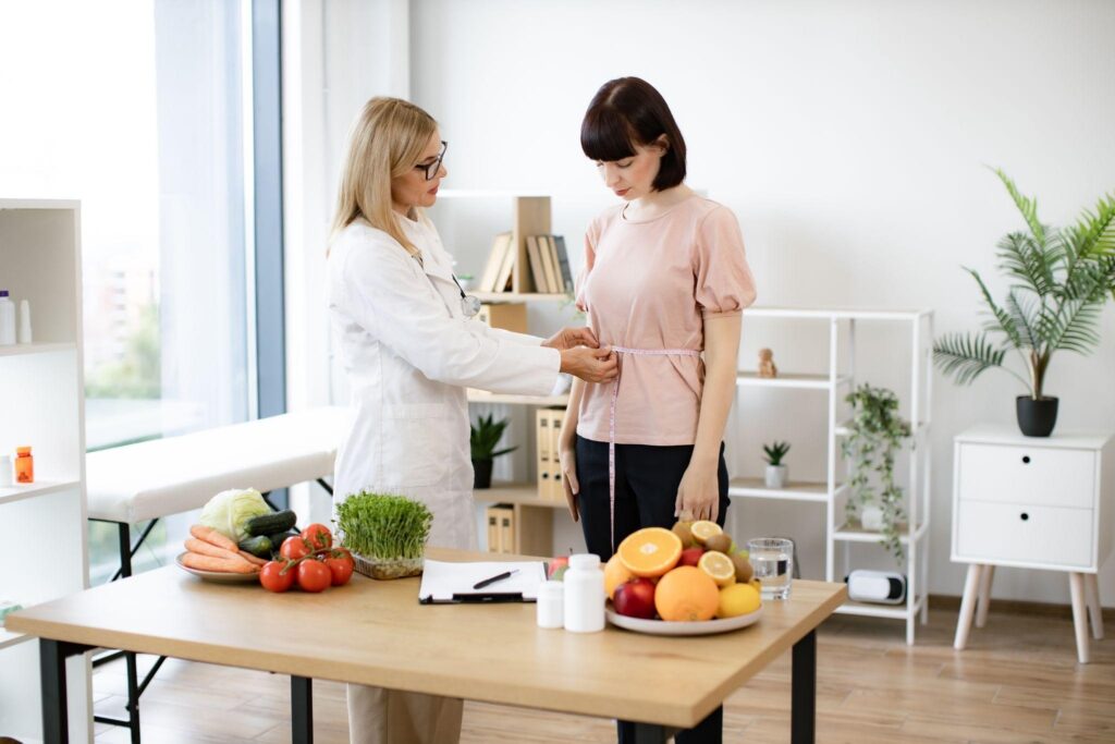 Weight loss doctor measuring woman’s waist at a wellness consultation with fruits and supplements on the table.