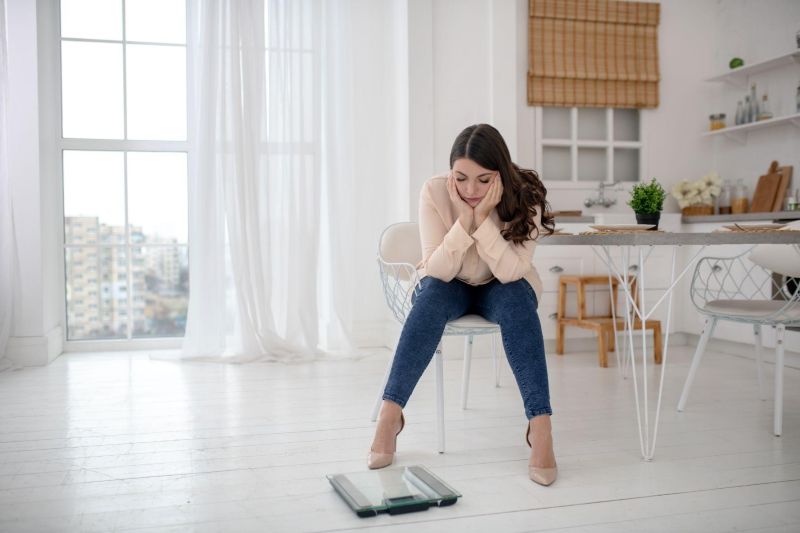 5 Signs Your Body Needs a Different Wilmington Weight Loss Approach Woman looking frustrated at a scale in her kitchen, struggling with weight loss despite efforts, a common concern in Wilmington weight loss journeys.