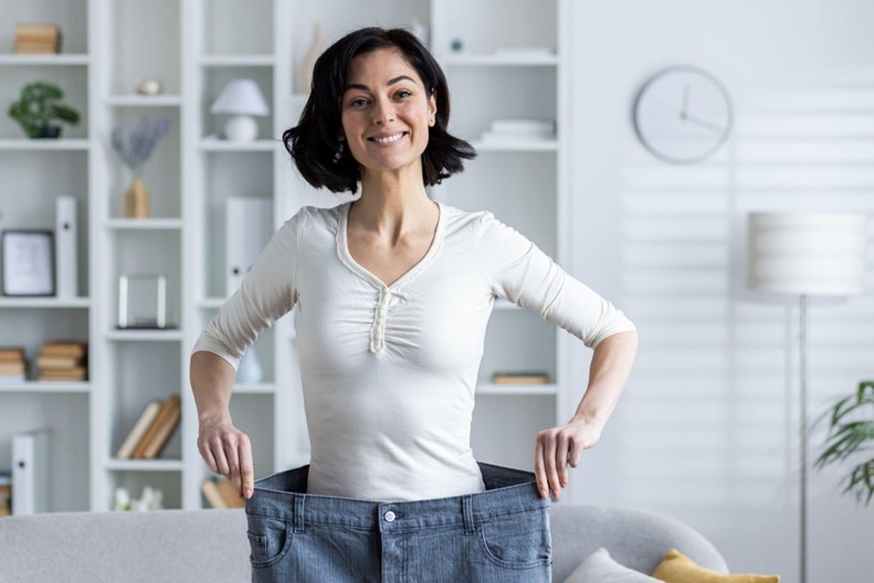 How Our Weight Loss Program Works With Your Body and Not Against It A young smiling woman standing at home and showing the result of dieting and losing weight.